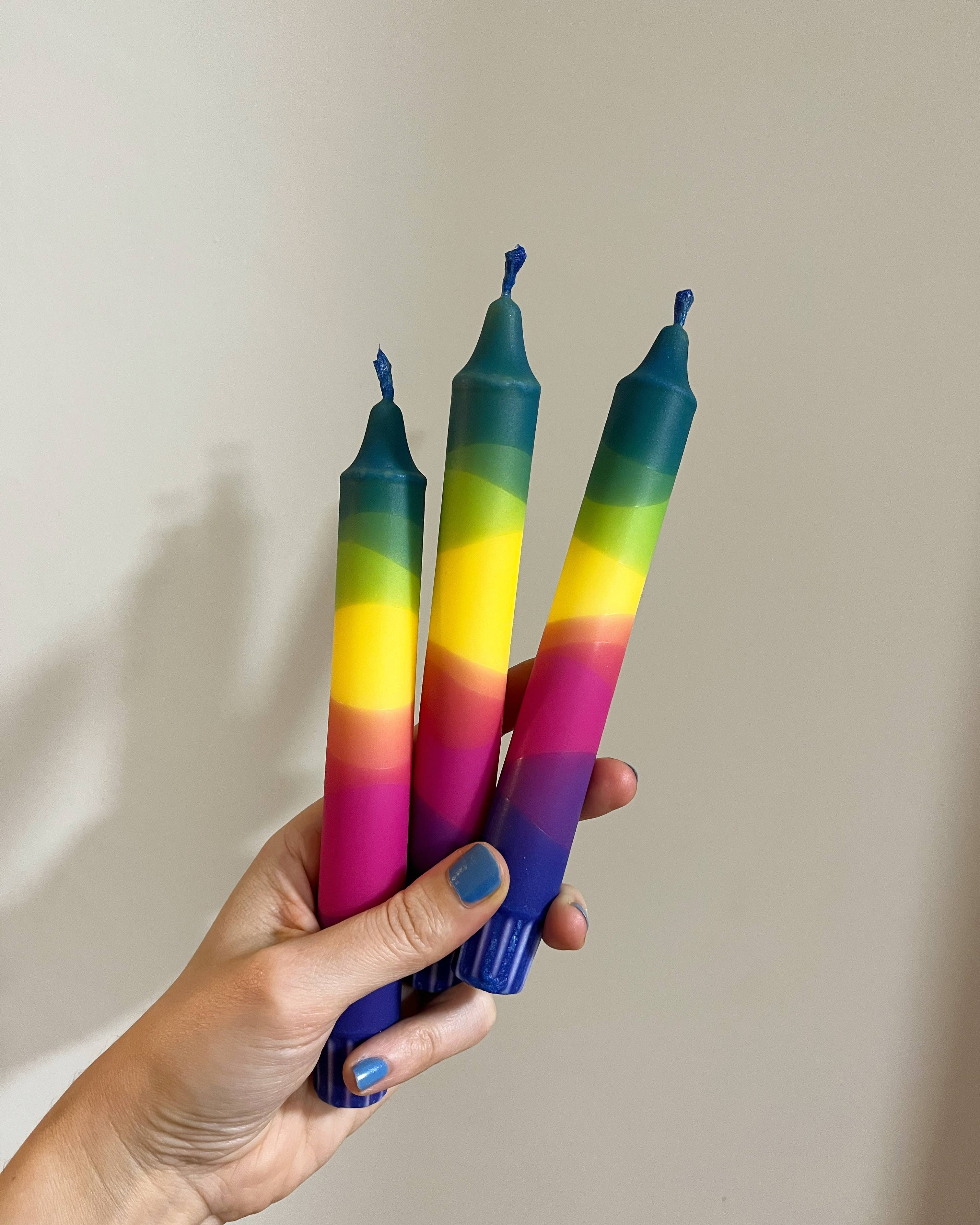 Hand holding three rainbow-colored candles against a plain background