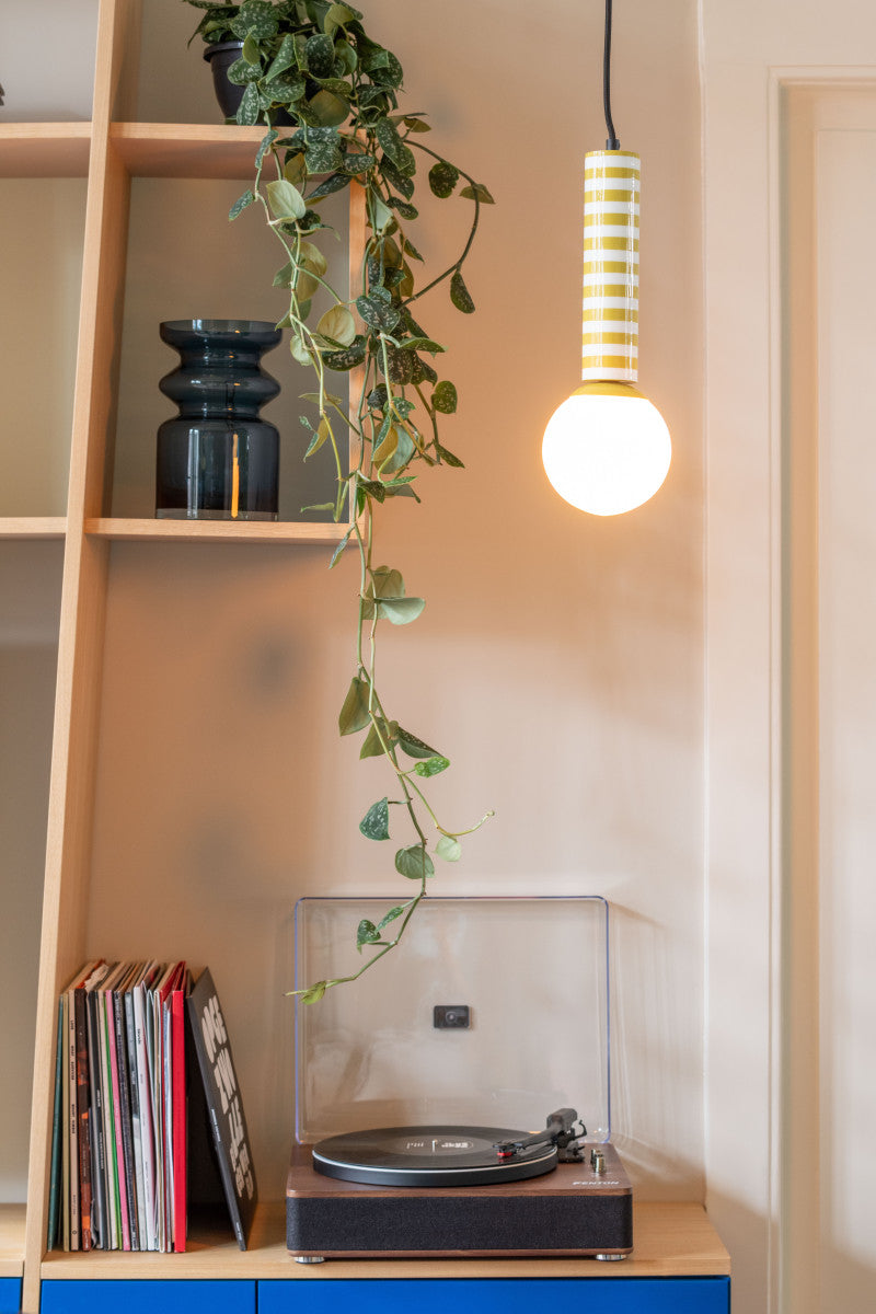 Record player on a wooden shelf with books and a plant, illuminated by a hanging light bulb.