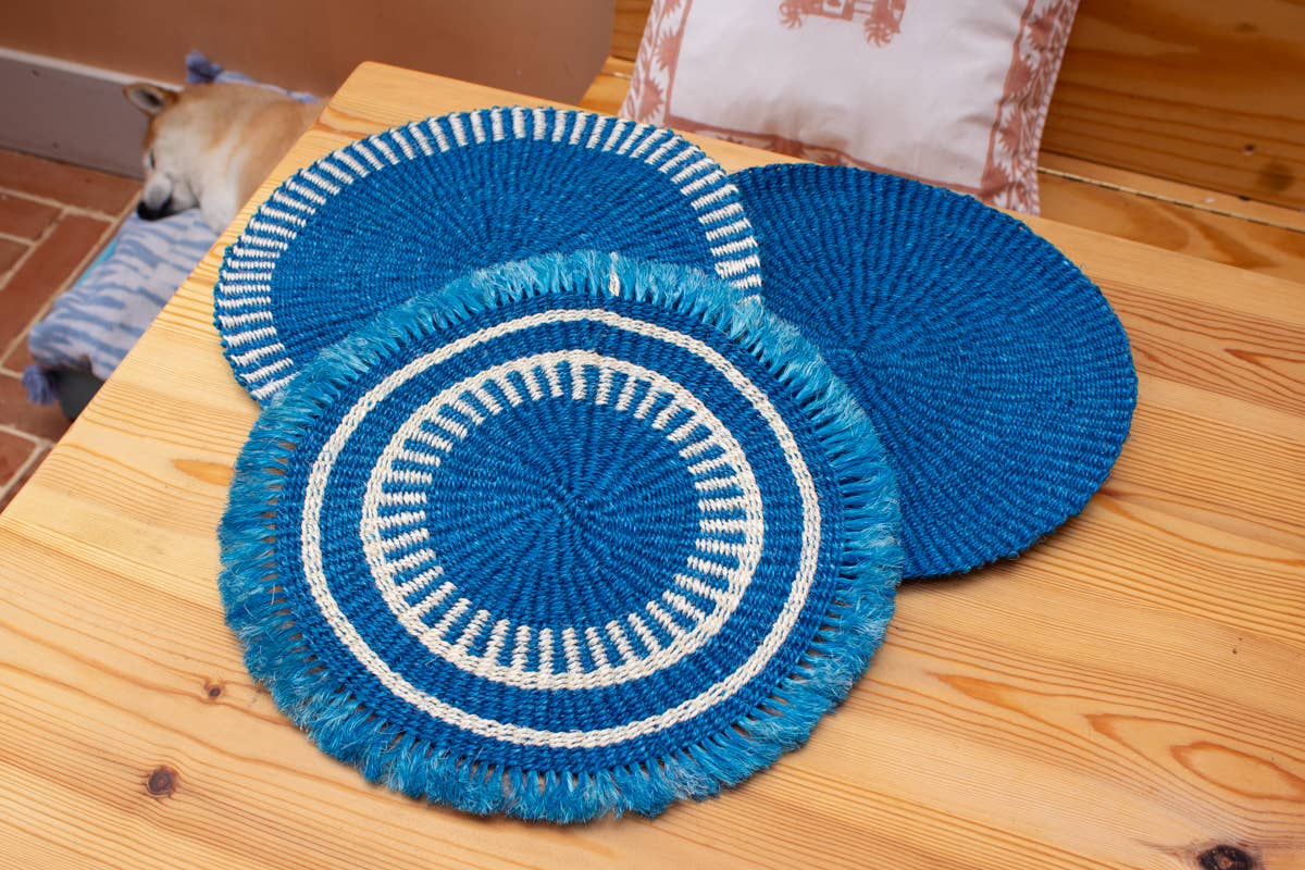 Set of blue and white woven placemats on a wooden table with a dog in the background.