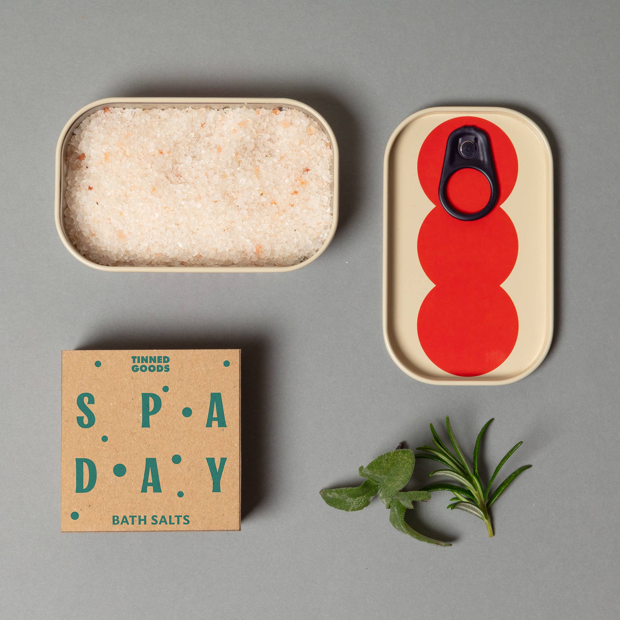 Tinned goods with a red lid, bath salts packaging, and green leaves on a gray background