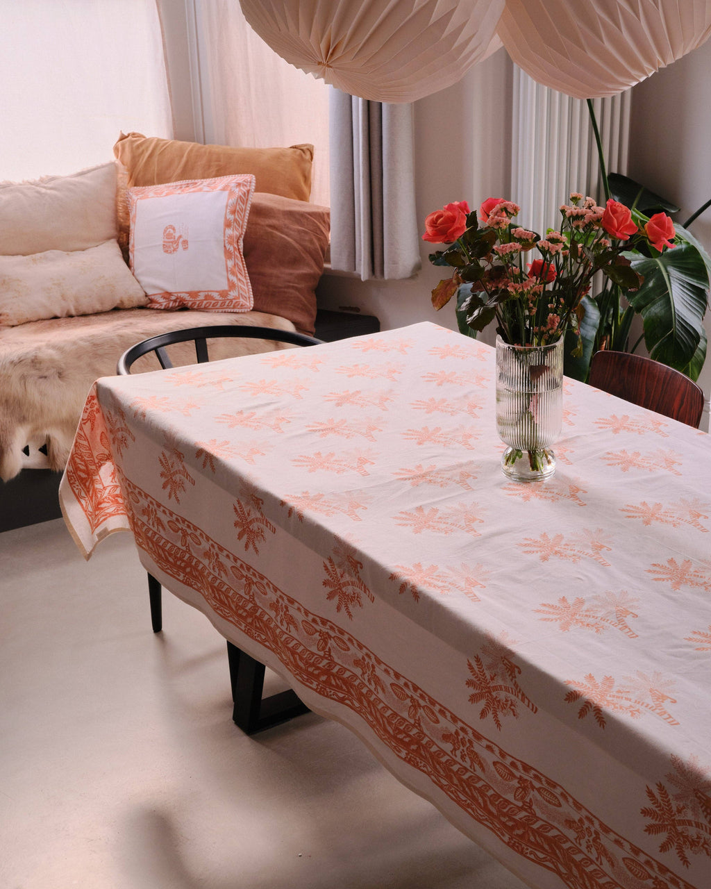 Living room with a table covered by an orange and white patterned tablecloth, flowers in a vase, and a couch.