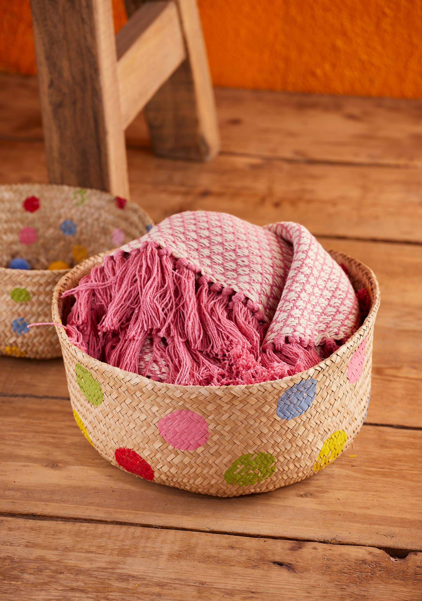 Woven basket with colorful polka dots containing a pink textured blanket on a wooden floor.