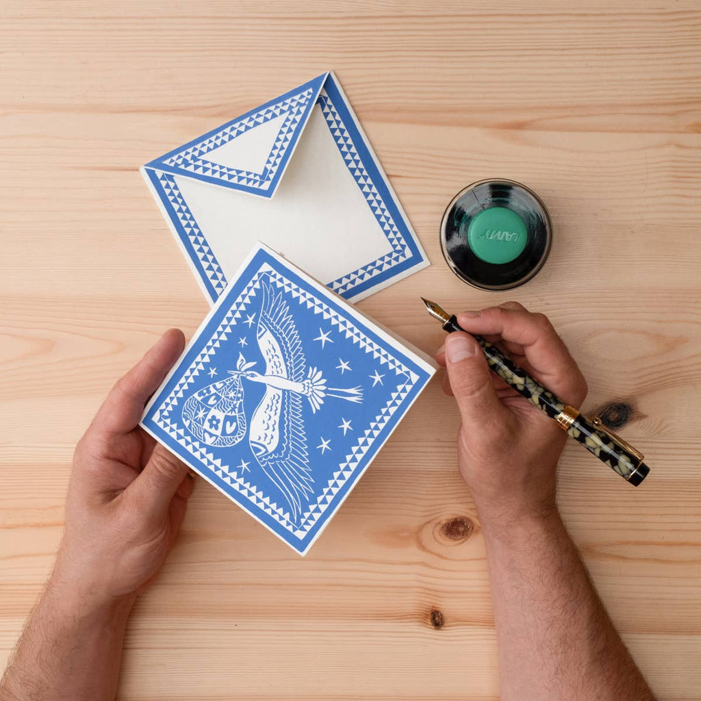 Person holding a blue card with white design on a wooden surface, next to a inkwell and pen.