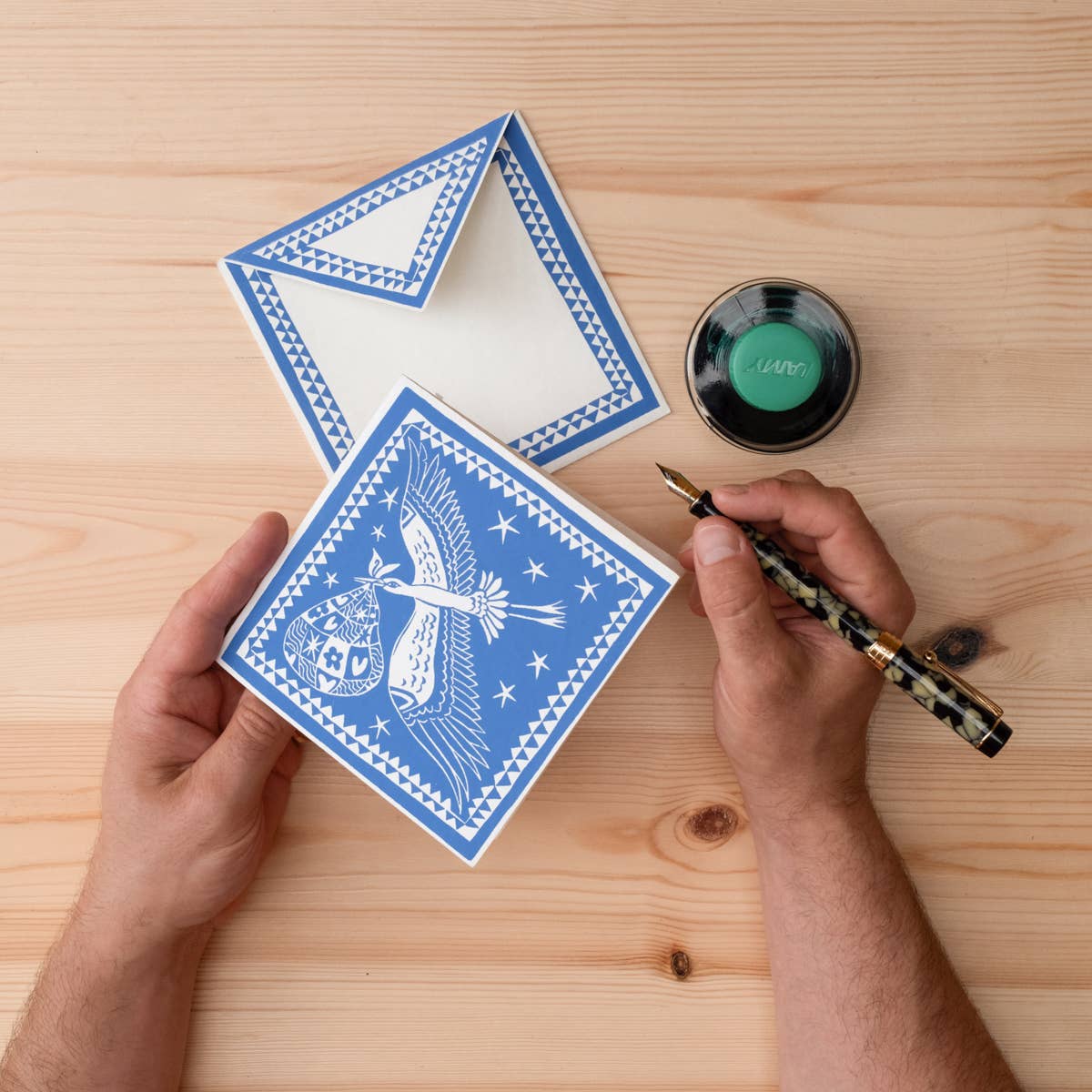 Person holding a blue card with white design on a wooden surface, next to a inkwell and pen.