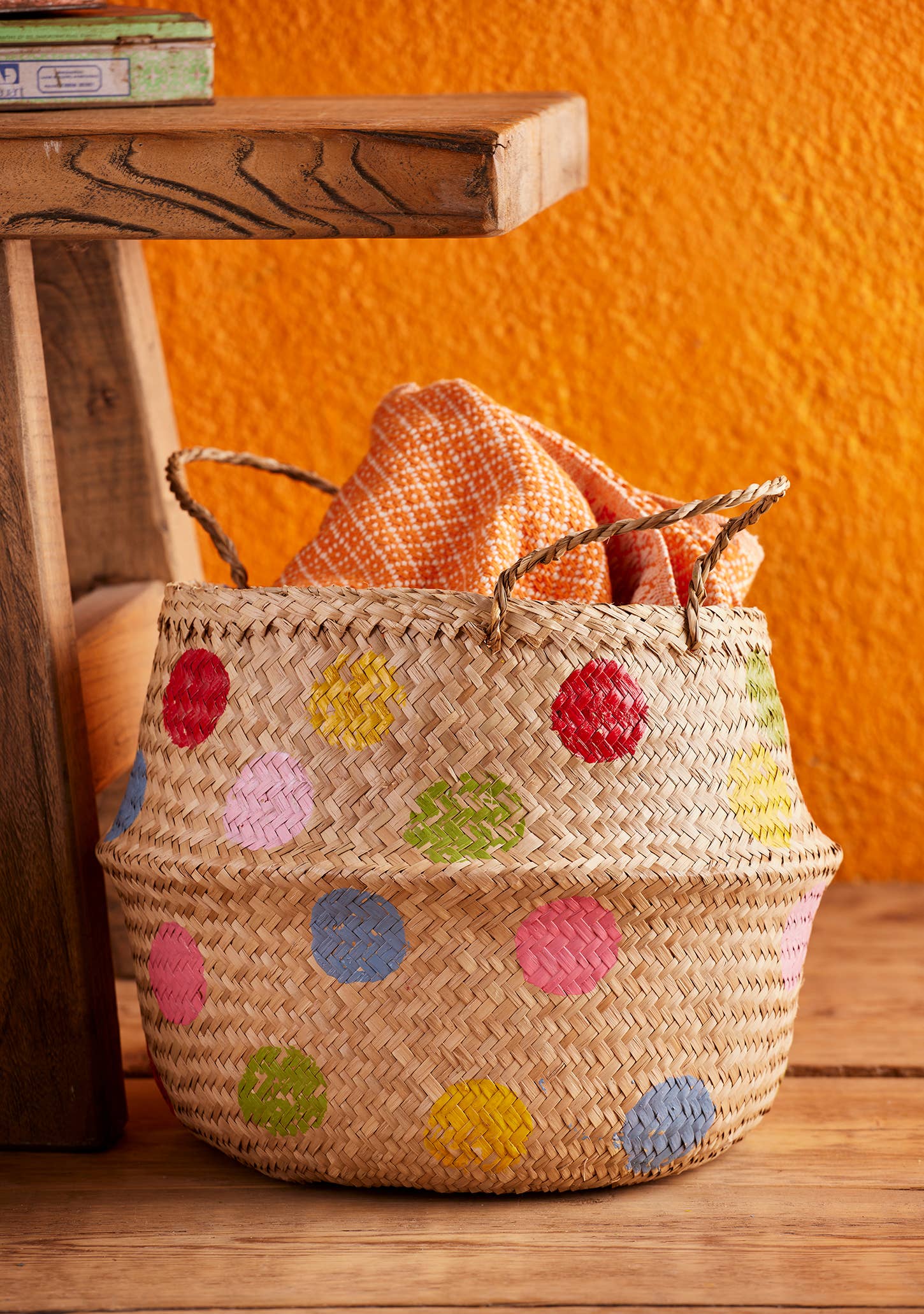Woven basket with colorful polka dots on a wooden surface against an orange wall.