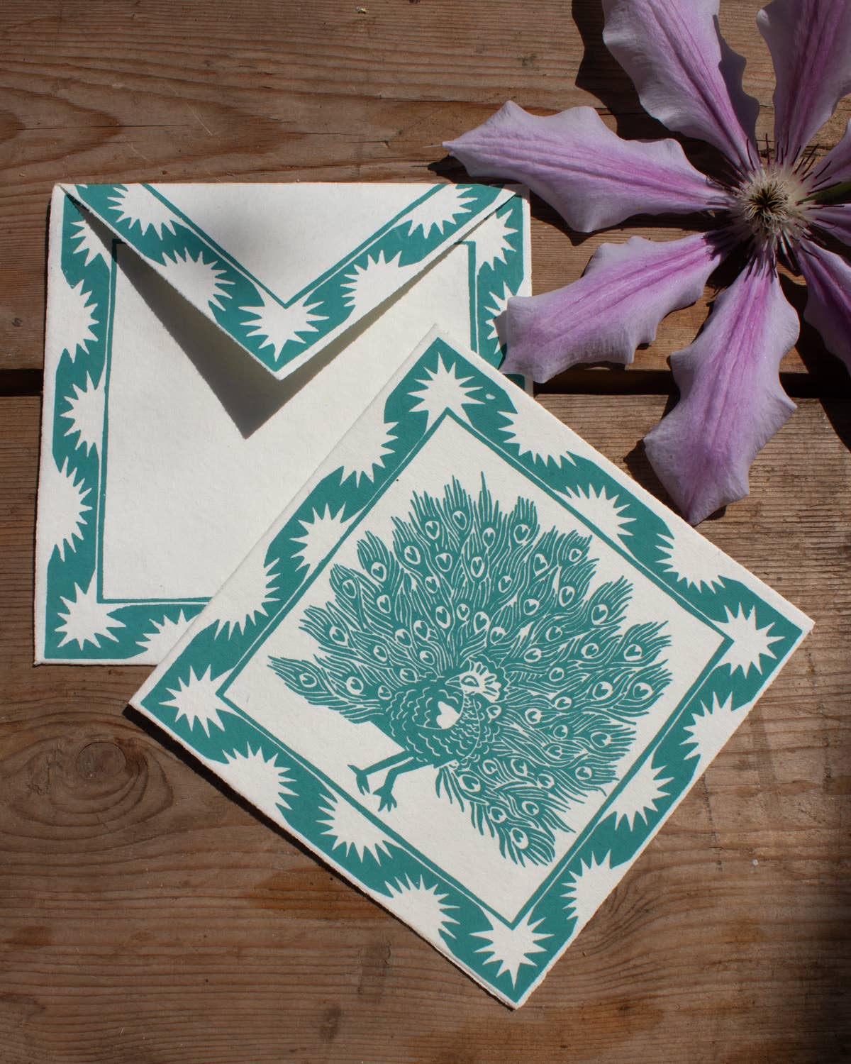 Two green and white peacock-themed cards on a wooden surface with a pink flower.