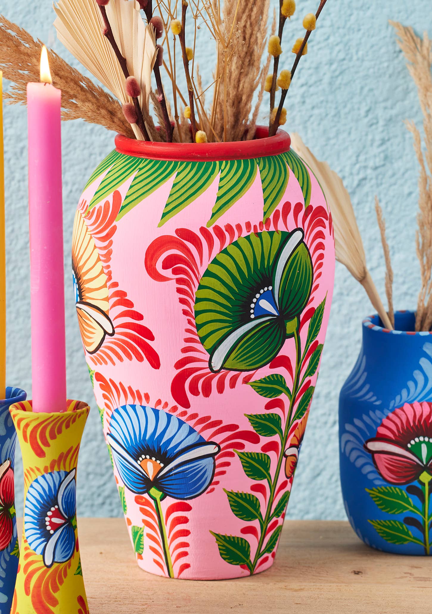 Colorful floral-patterned vase with a pink candle and dried plants on a wooden surface.