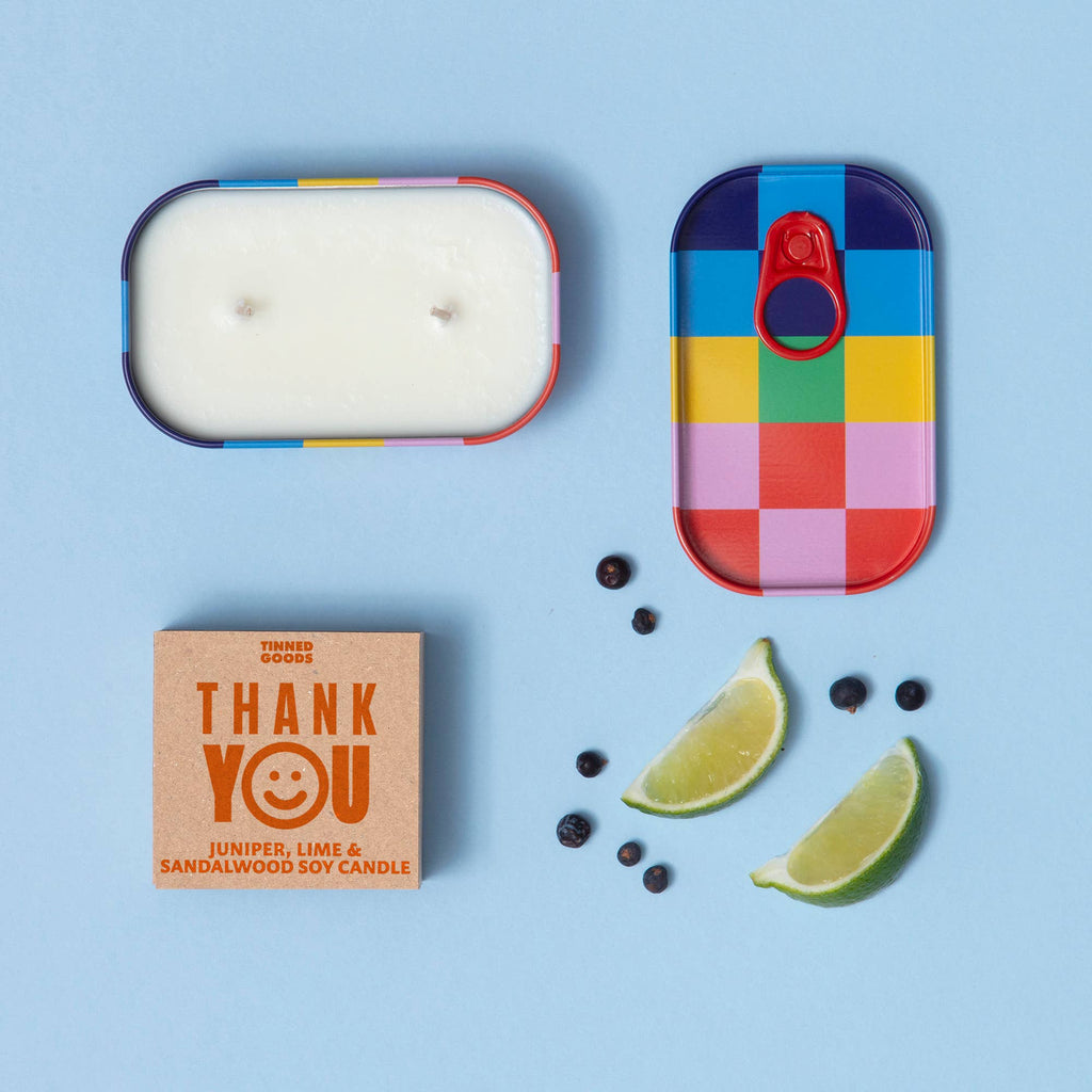 Colorful candle tin with a candle inside, a small box labeled 'THANK YOU', lime slices, and berries on a blue background.