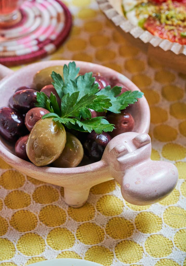 Small pink hippo dish with olives and parsley on a yellow patterned tablecloth