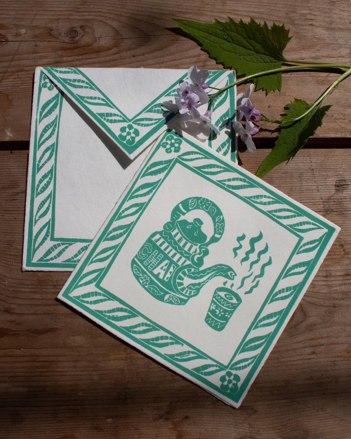 Two green and white patterned cards on a wooden surface with a leaf and flowers.