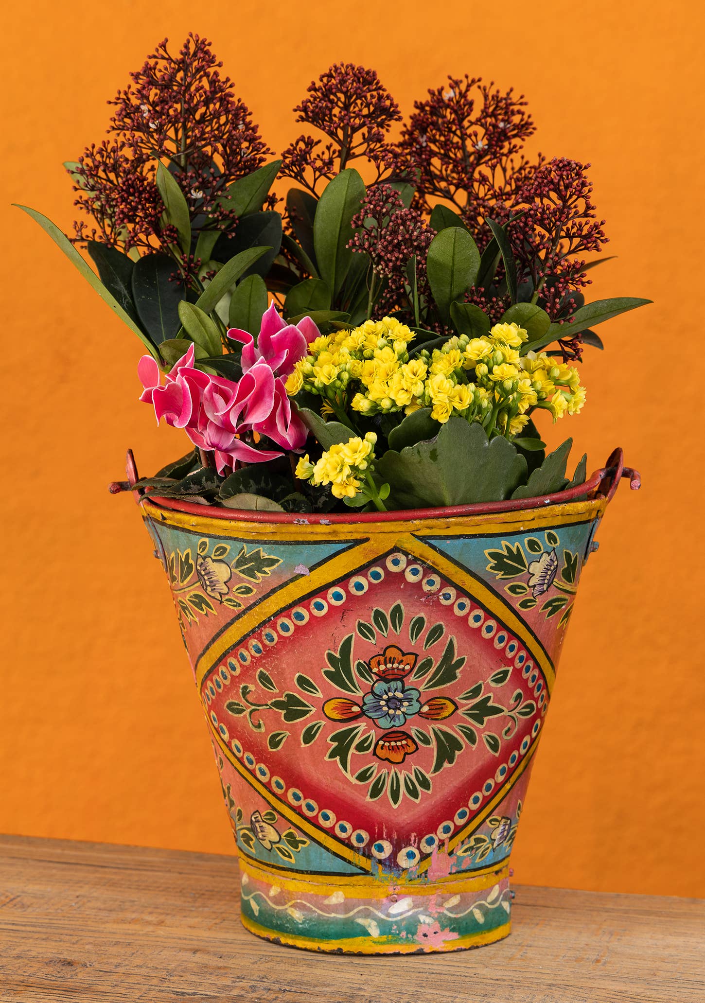 Decorative bucket with colorful patterns filled with flowers on a wooden surface and orange background
