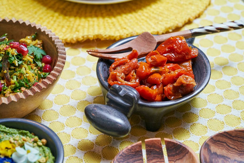 Dish of roasted red peppers with a wooden spoon on a yellow patterned tablecloth.