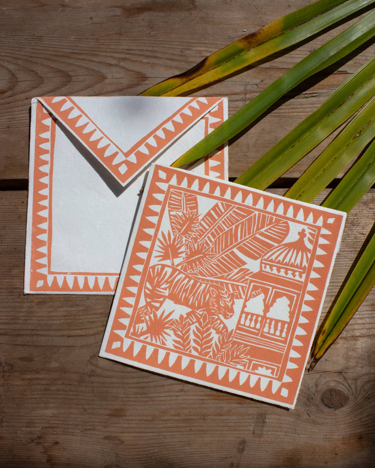 Two decorative cards with orange patterns on a wooden surface with palm leaves.
