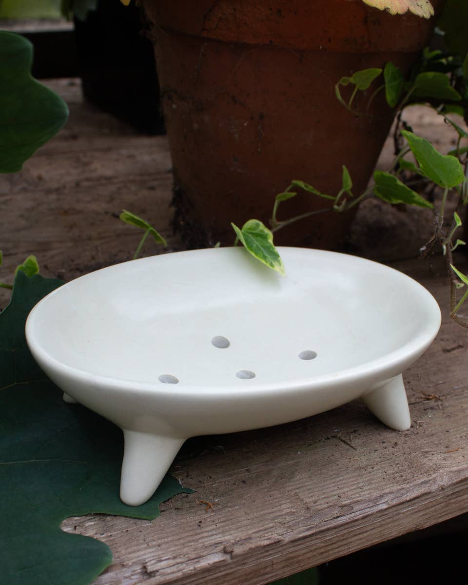 White soapstone soap dish with three legs on a wooden surface with leaves in the background