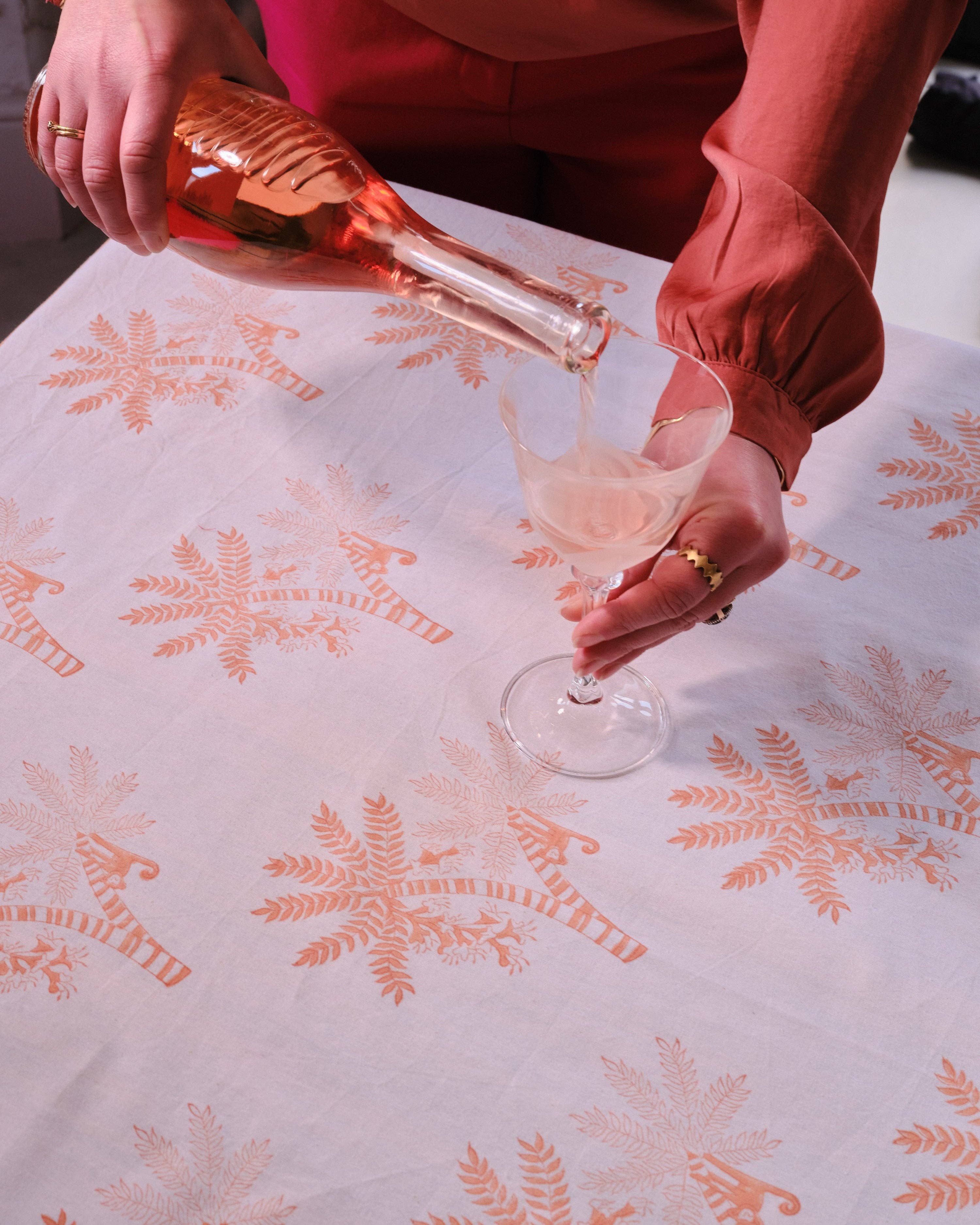 Person pouring pink wine into a glass on a tablecloth with orange palm tree  patterns