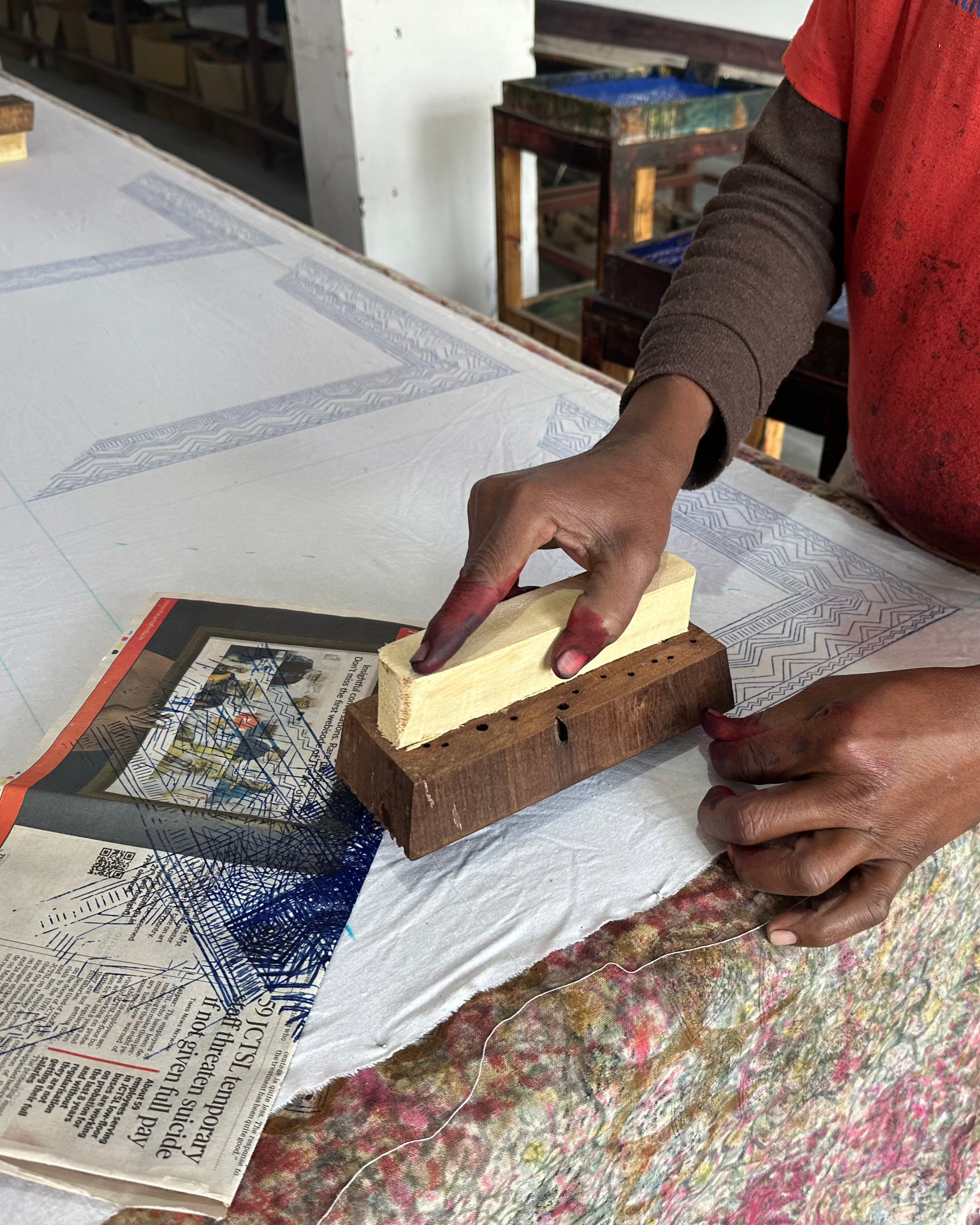 Person using a printing block with a wooden handle on a table with newspapers and patterned fabric.